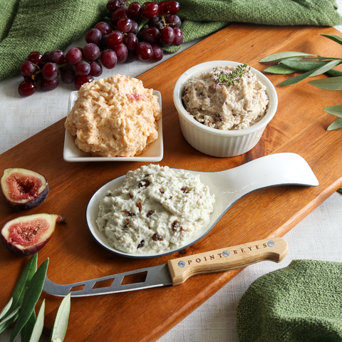 Product: Three bowls of different types of cheese on a wooden board with figs and grapes, featuring a Point Reyes knife.
