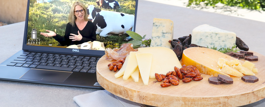 a cheeseboard in front of a laptop showing a woman hosting a virtual cheese tasting