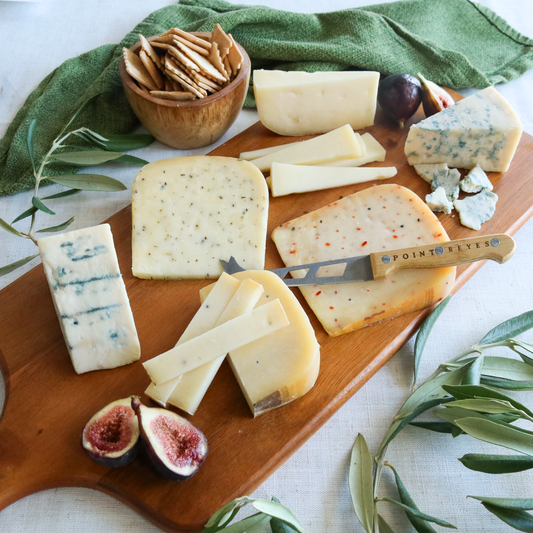 Assorted cheeses on a wooden board with figs and crackers, surrounded by greenery.