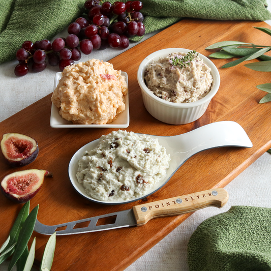Three bowls of different types of cheese on a wooden board with figs and grapes, featuring a Point Reyes knife.