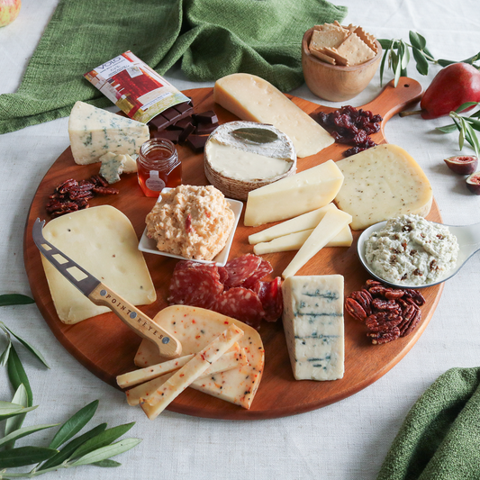 Assorted cheeses, meats, and accompaniments on a wooden board with a green cloth and fruit in the background.